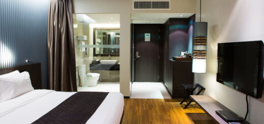 Hotel room with a large bed, dark headboard, white linens and a brown runner; a wall TV faces the bed, bathroom visible nearby.