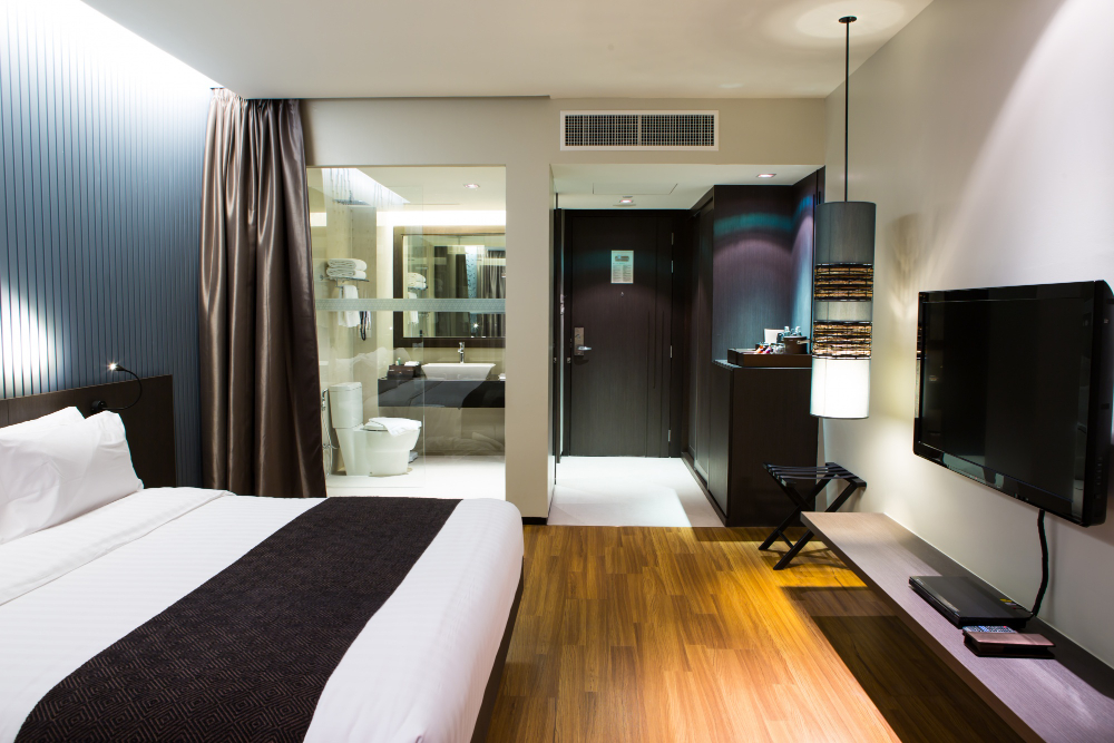 Hotel room with a large bed, dark headboard, white linens and a brown runner; a wall TV faces the bed, bathroom visible nearby.