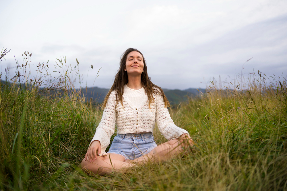 Woman sits cross‑legged in a grassy field with eyes closed, practicing meditation in a light cardigan and denim shorts under a cloudy sky