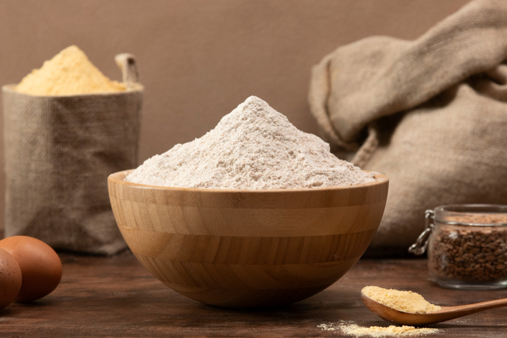 Wooden bowl piled with flour, with eggs and sacks of ingredients in the background for baking.