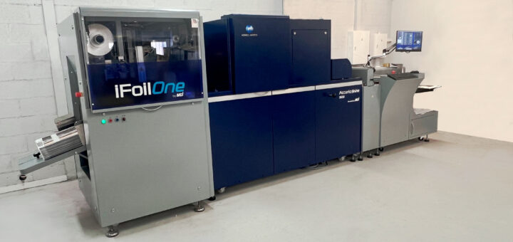 Industrial printing/foil stamping machine (iFoilOne) in a clean room, blue and gray cabinet setup for foil application on sheets; control panel visible on left side.