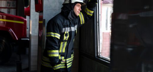 Firefighter in full gear leaning against a doorway, looking out a window from inside a fire station.