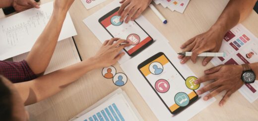 Team members collaboratively review a mobile app prototype, pointing at colorful screen mockups on a desk with papers and icons nearby.