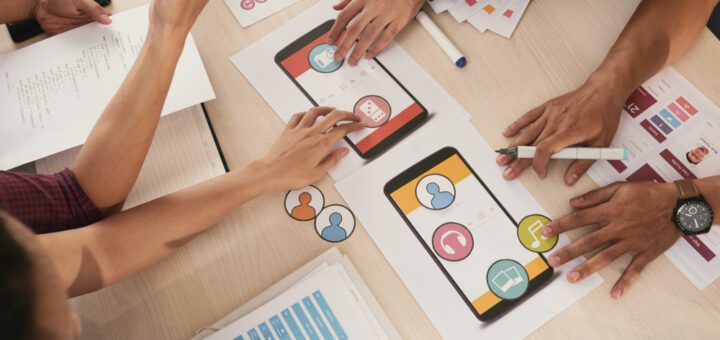 Team members collaboratively review a mobile app prototype, pointing at colorful screen mockups on a desk with papers and icons nearby.