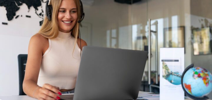 Woman wearing a headset and smiling at a laptop on a white desk with a world map wall behind her.