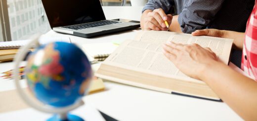 Two people studying at a desk with a laptop, an open book, and a yellow pencil nearby by a windowed wall.