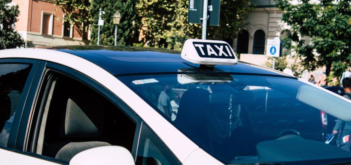 White taxi car with a rooftop TAXI sign parked on a sunny city street; driver visible through the windshield.