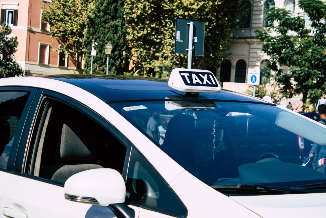 White taxi car with a rooftop TAXI sign parked on a sunny city street; driver visible through the windshield.