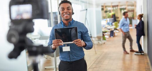 Smiling man wearing a headset holds a tablet while filming with a camera in an office setting.