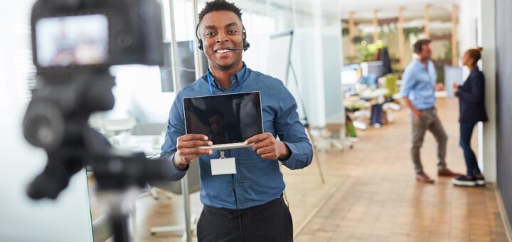 Smiling man wearing a headset holds a tablet while filming with a camera in an office setting.