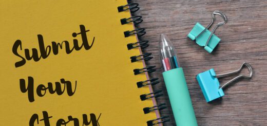 Yellow notebook with 'Submit Your Story' beside a teal pen and binder clips on a wooden desk.