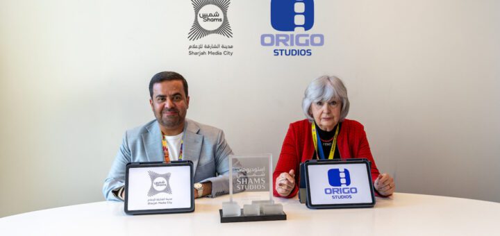 Two people sit at a white table displaying tablets with Shams and Origo Studios logos; backdrop shows both logos.