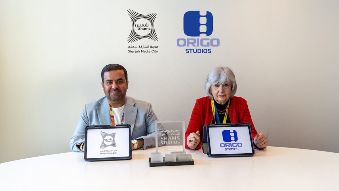 Sharjah y Origo - IP | ComunicaTech.Com Two people sit at a white table displaying tablets with Shams and Origo Studios logos; backdrop shows both logos.